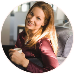 A portrait, enclosed in a circular frame, of a beautiful, smiling woman with long brown hair, seated and holding a white mug. She is wearing a maroon or dark red satin blouse and is looking confidently at the camera. Sunlight illuminates her hair from the side.