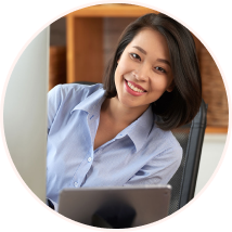 A headshot, enclosed in a circular frame, of a smiling young Asian woman with short dark hair, wearing a light blue collared shirt. She is looking directly at the camera, leaning slightly around a white partition or monitor, with a tablet visible in the foreground. The background is an office setting.