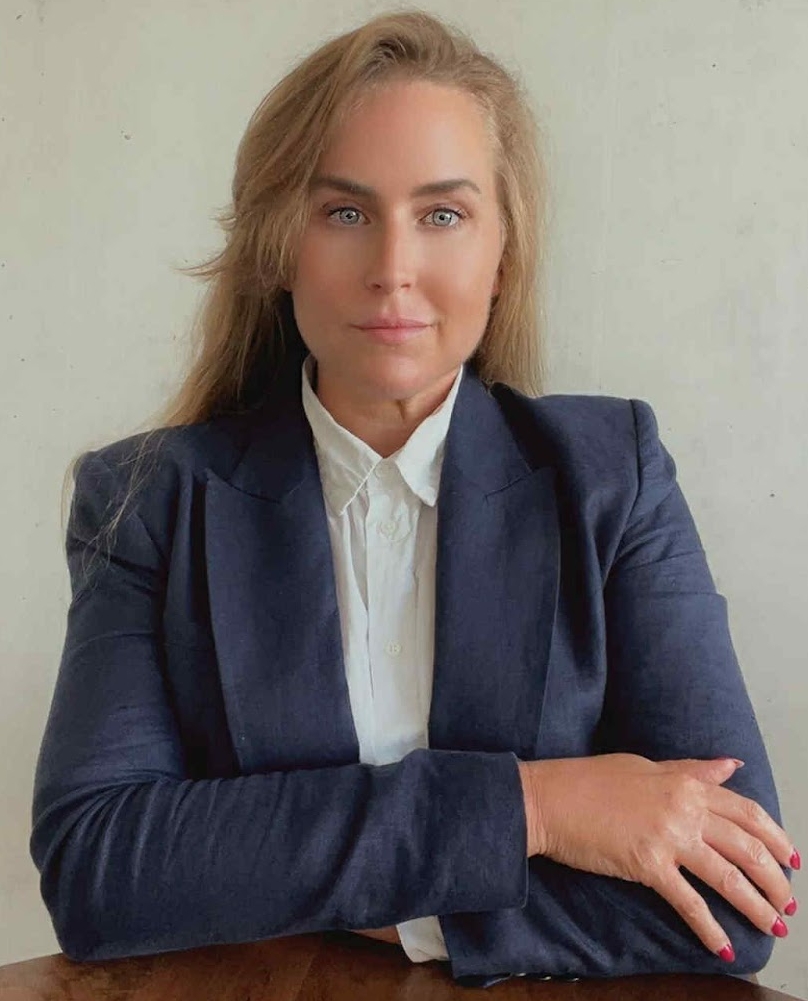 Professional headshot of a woman with long blonde hair, wearing a dark navy blazer and a white shirt, looking directly at the camera with her hands clasped on a table.